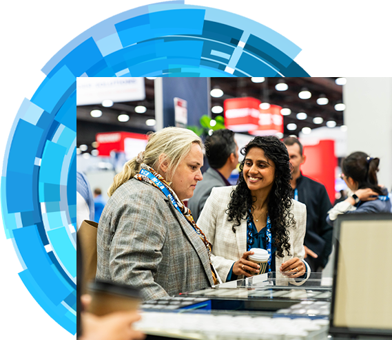 Two women engaged in conversation at a bustling trade show, surrounded by various booths and attendees